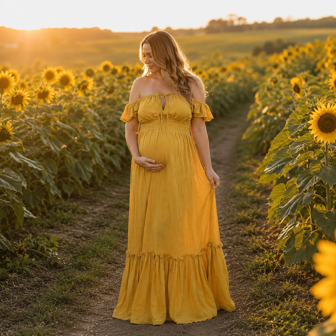 A pregnant woman in a yellow off-shoulder gown stands on a sunflower field path; the dress has ruffled sleeves, a front tie, elasticated waist, and tiered ruffled hem. 