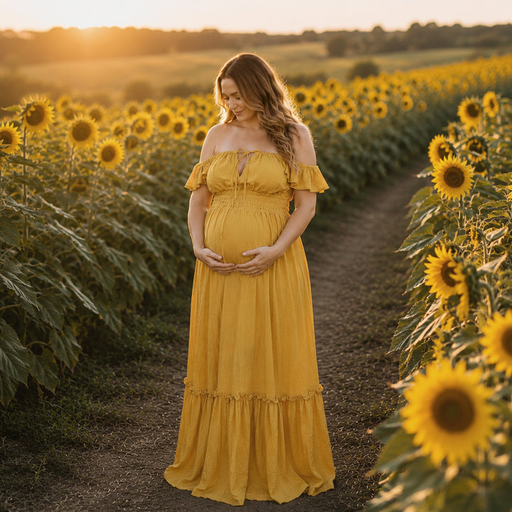 A pregnant woman in a yellow off-shoulder gown stands on a sunflower field path; the dress has ruffled sleeves, a front tie, elasticated waist, and tiered ruffled hem. 