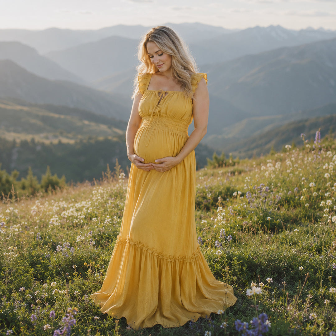 A pregnant woman in a yellow square-neck gown stands on a mountain meadow; the dress has ruffled straps, a front tie, elasticated waist, and tiered ruffled hem. 