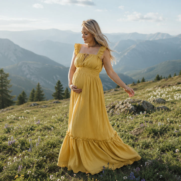A pregnant woman in a yellow square-neck gown stands on a mountain meadow; the dress has ruffled straps, a front tie, elasticated waist, and tiered ruffled hem. 