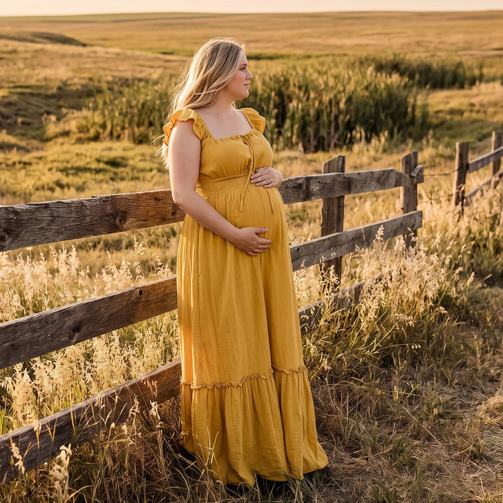 A pregnant woman in a yellow square-neck gown stands sideways by a wooden fence; the dress has ruffled straps, a front tie, elasticated waist, and tiered ruffled hem. 