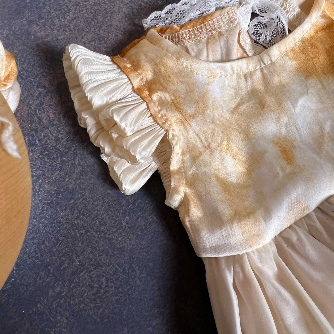 A close-up of a yellow tie-dye baby romper dress, showing the round neck, white lace and one ruffled sleeve, laid on a dark surface.