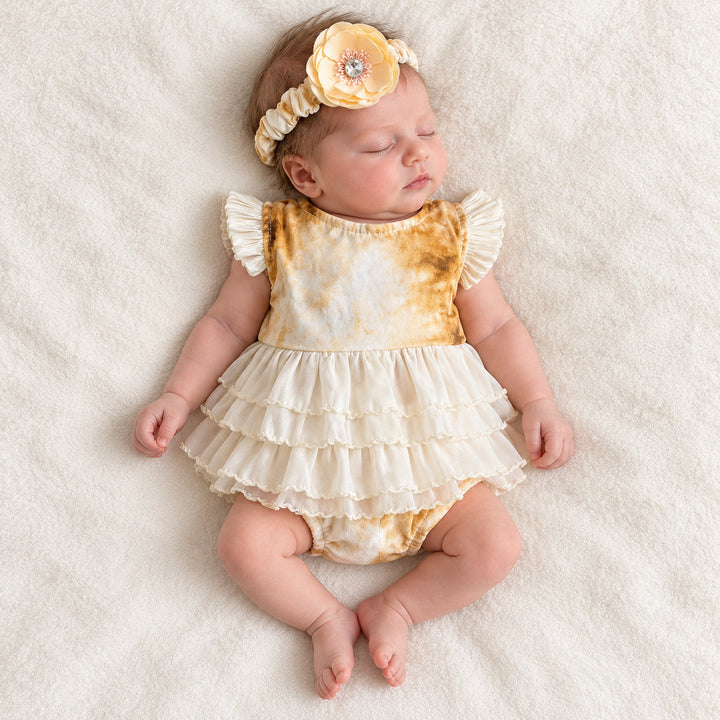 A baby in the yellow tie-dye romper and flower headband lies on her back on a white blanket, eyes closed.