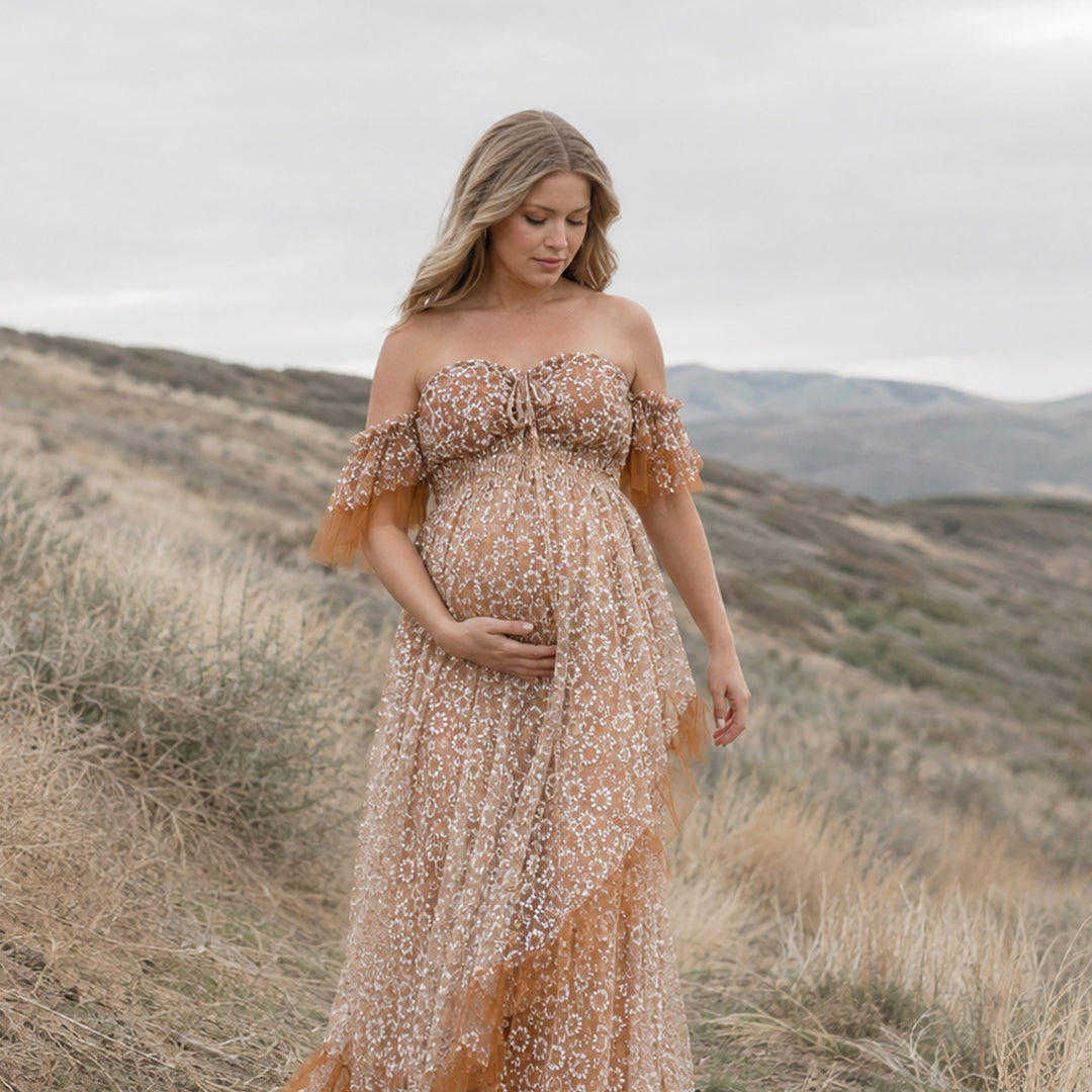 A  pregnant woman is wearing Khaki Tyche stands on hills and dry vegetation; she looks down, cradling her belly with her right hand.