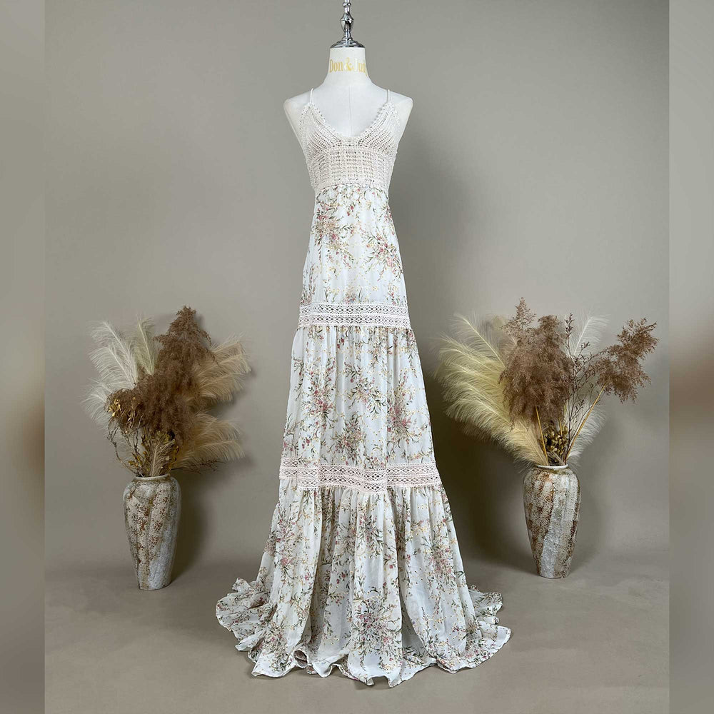 The Urania,an apricot bottom floral maternity maxi dress with spaghetti straps, a crochet bodice, and lace-trimmed tiered panels on the skirt, displayed on a mannequin. The dress features a V-neckline and a floor length hem with ruffled edges.