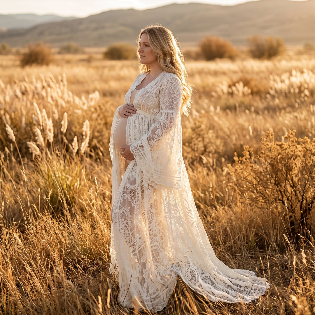 A pregnant woman stands on grass wearing a beige lace maternity gown with long bell sleeves, an open front, and a train that spreads across the ground.
