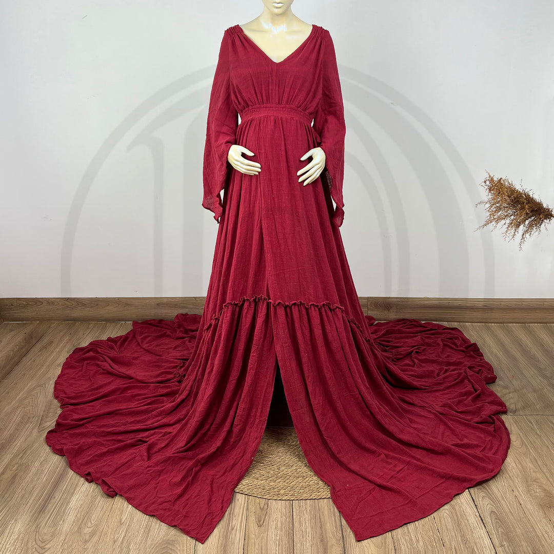 A Daphne bordeaux long-sleeved V-neck gown is displayed on a mannequin, with hands positioned on the simulated belly. The dress features a high front slit and its hem and train are spread out on the wooden floor.