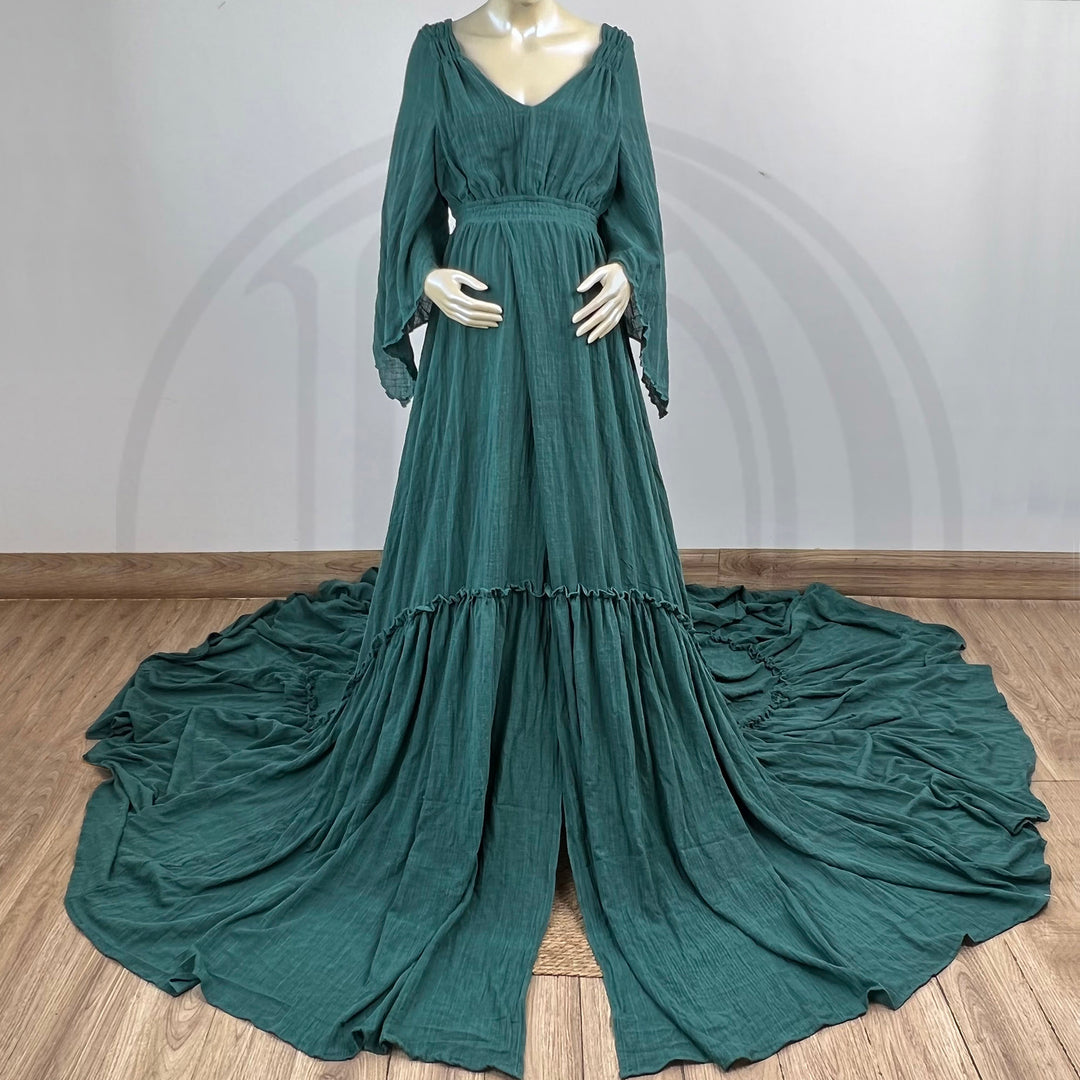 A Daphne emerald long-sleeved V-neck gown is displayed on a mannequin, with hands positioned on the simulated belly. The dress features a high front slit and its hem and train are spread out on the wooden floor.