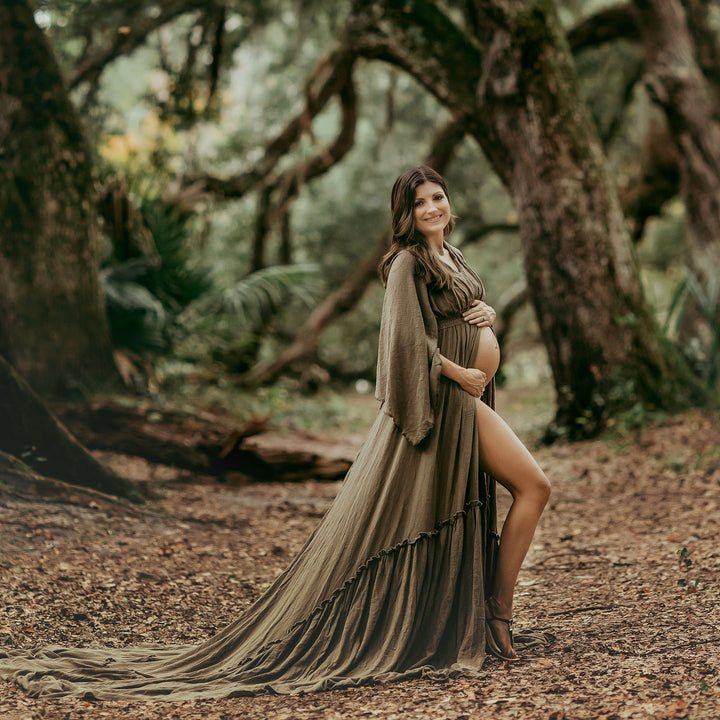 a pregnant woman in a Daphne olive long dress with a slit and shawl stands on leaf-covered forest ground, holding her belly.