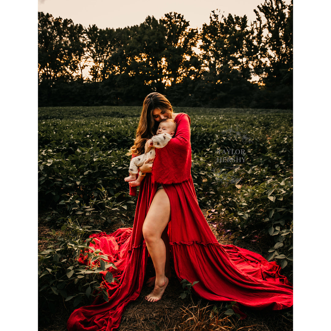 a woman in a Daphne red slit dress stands barefoot among plants, holding an infant. A text watermark appears on the right.